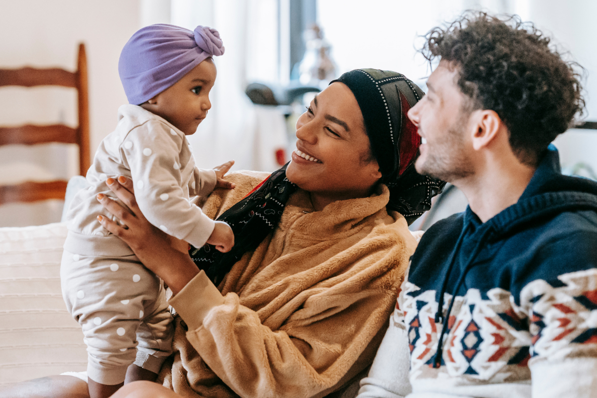 Two parents, one with dark brown skin and one with pale skin, smile with their infant, who is wearing an adorable purple headwrap.