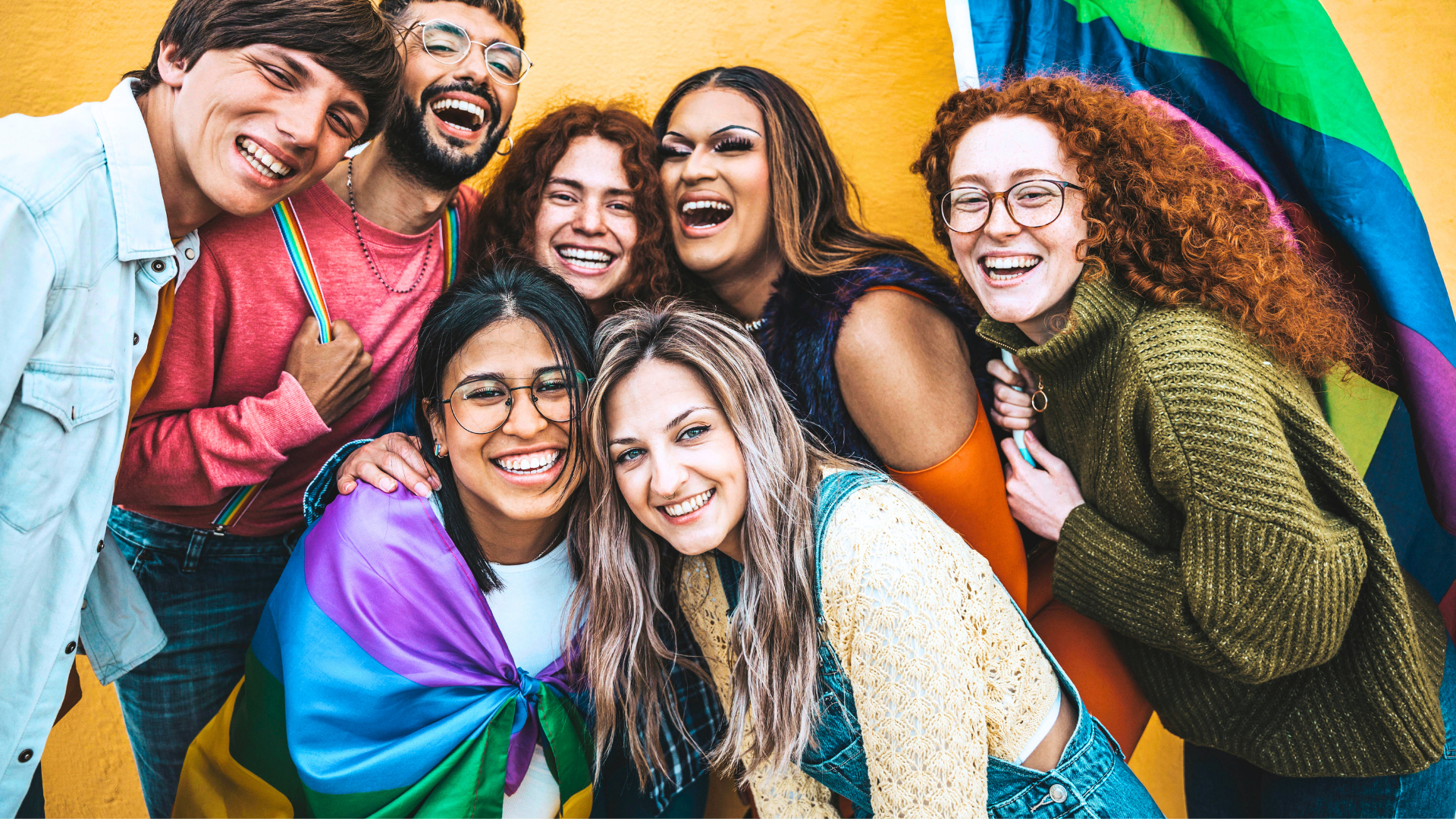A group of smiling, diverse individuals with pride flags wrapped around two of the members of the group. 