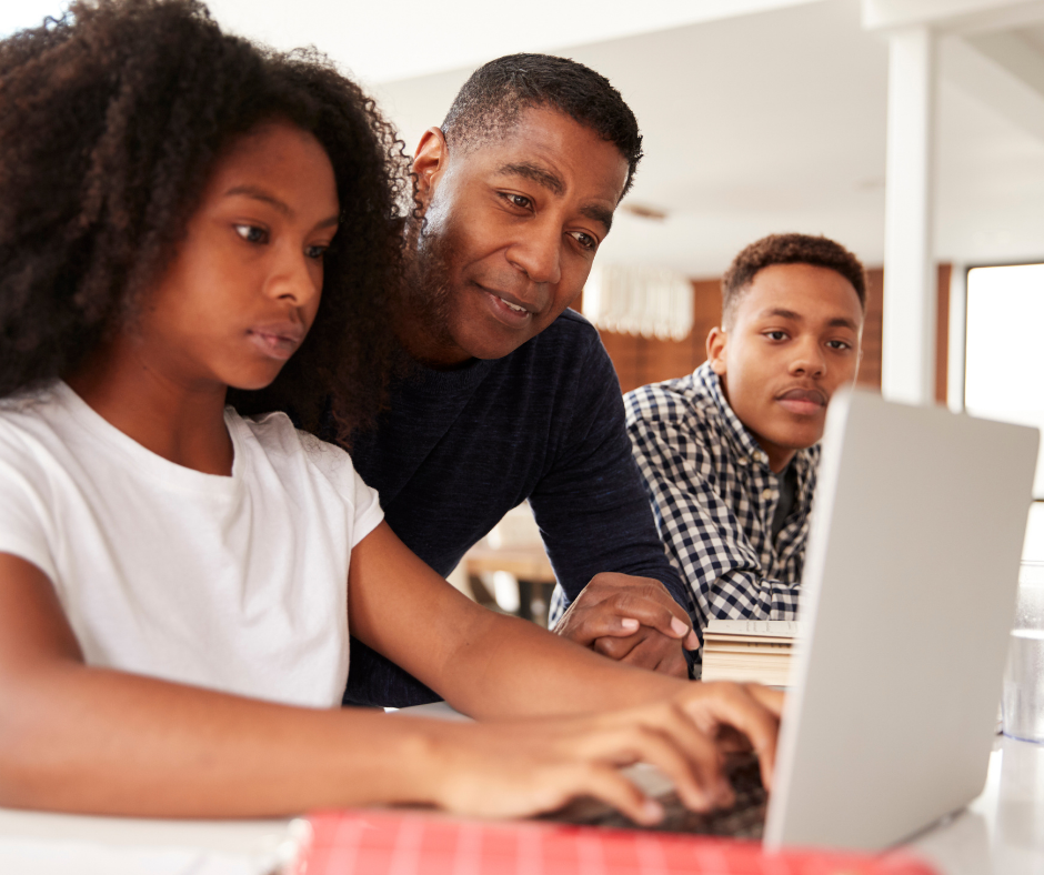 A Black teen girl types on a laptop at her kitchen counter. Her father sits beside her attentively, leaning in to see what she is looking at while her teen brother watches.