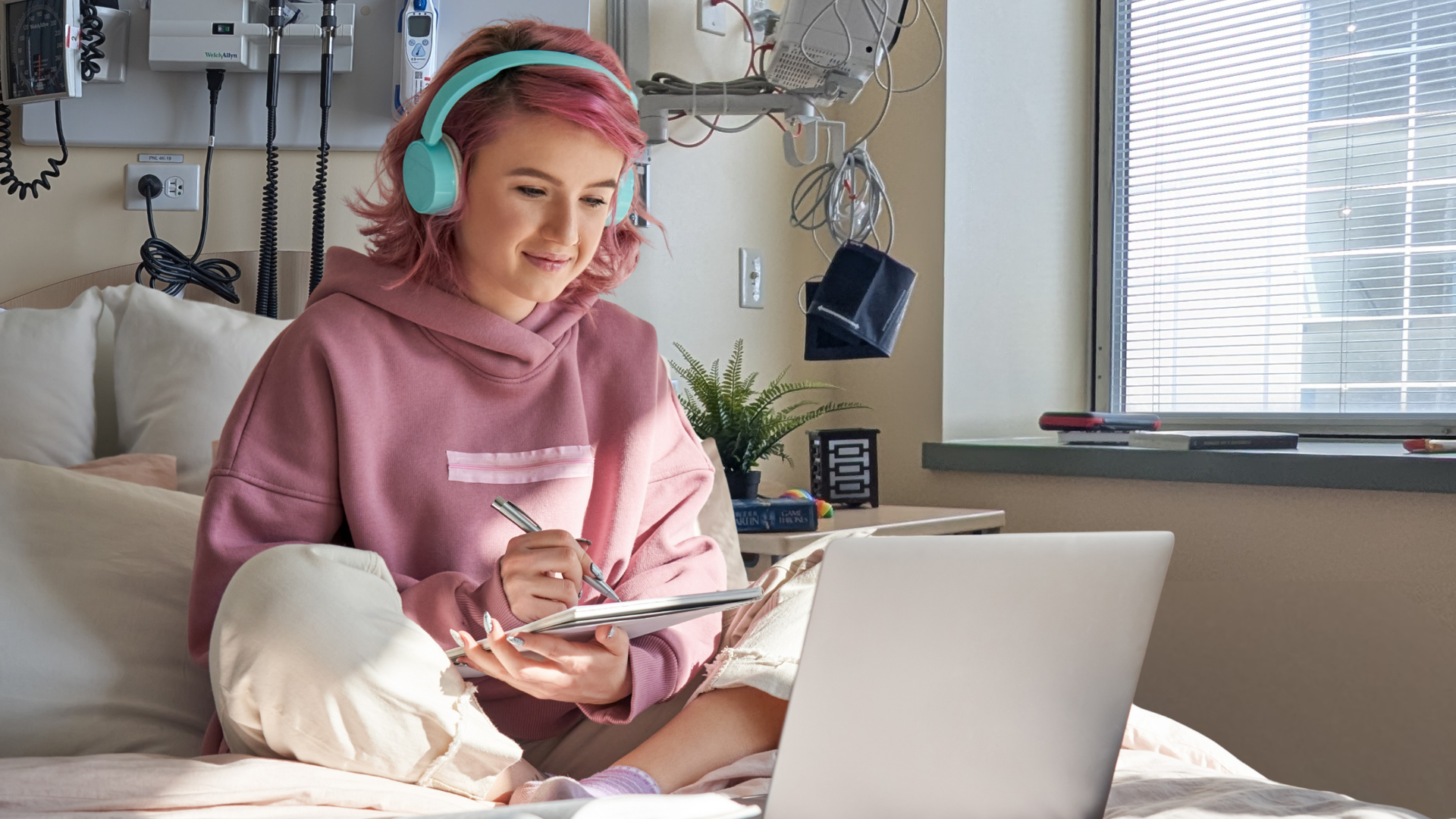 A young caucasian woman with pink hair and a pink hoodie sits on a bed taking notes as she watches something on her laptop. Medical equipment is visible on the wall behind the bed, as in a hospital room.