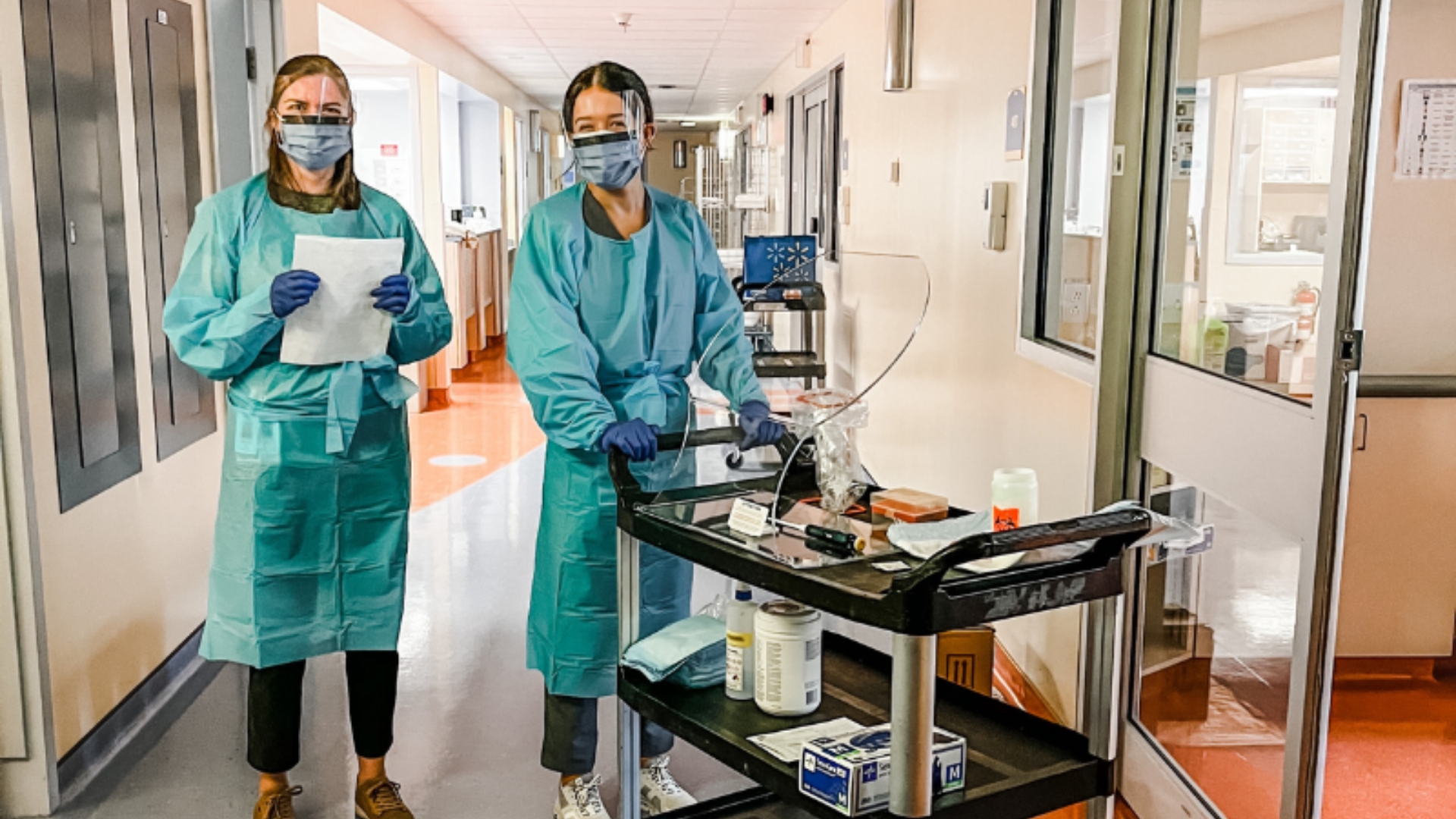 Two CCfV nurses stand in full Personal Protective Equipment outside a participant's room with a cart of medical materials, ready to begin their rounds for the day. 