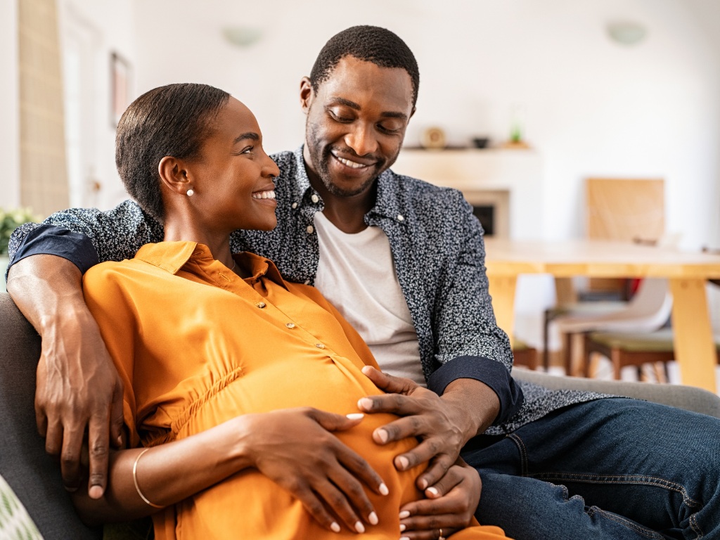 A young Black couple (a man and a woman) smile at each other while sitting on their sofa. They both have a hand placed affectionately on the woman's stomach - she is pregnant.