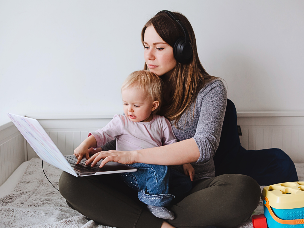 A white woman types on a laptop, seated cross-legged on the floor with an infant sitting on her lap.