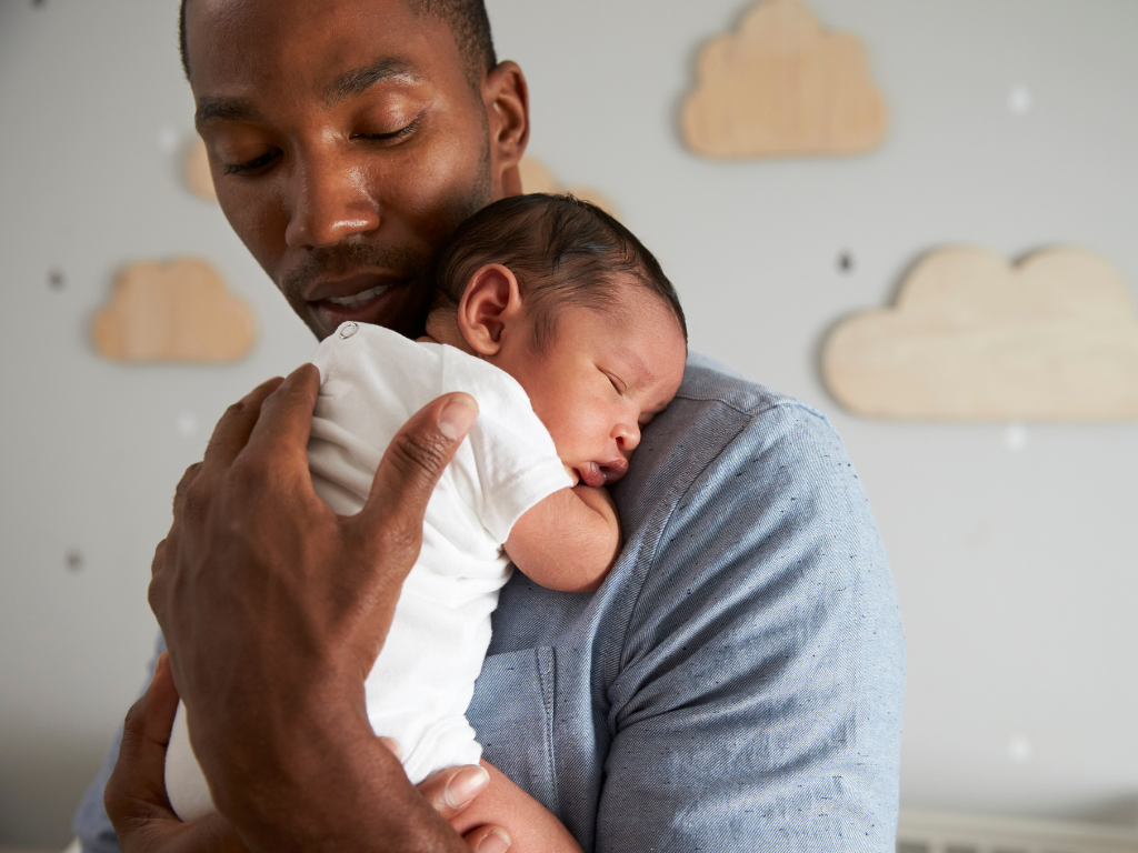A Black father holds his sleeping newborn on his shoulder, looking at them with care. The wall of a nursery with cloud decals is visible in the background.