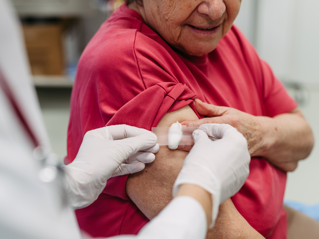 An elderly woman with light skin watches as a pharmacist tapes a cotton ball on her arm where she's just received a vaccine.