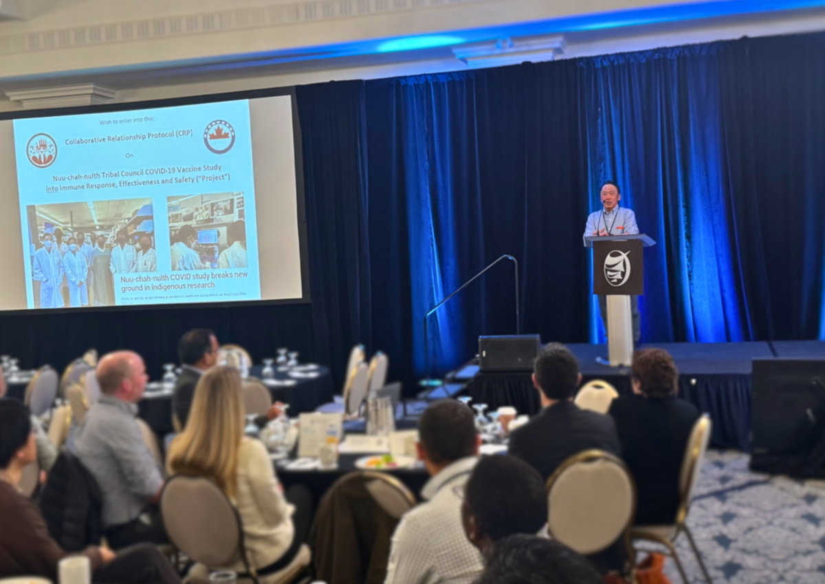  Dr. John Kim gives a lecture to a room. A powerpoint presentation is shown to the left-hand side and there are people watching at tables.