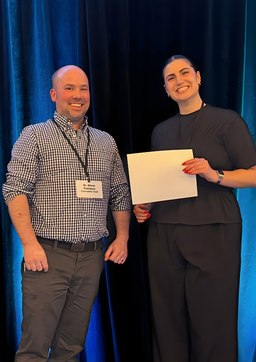 Dr.Glenn Patriquin wearing a plaid shirt stands next to Bailey Selig wearing all black. Bailey holds the certificate for Best Post Presentation - Research Associate, though the printed side is facing away from the audience.
