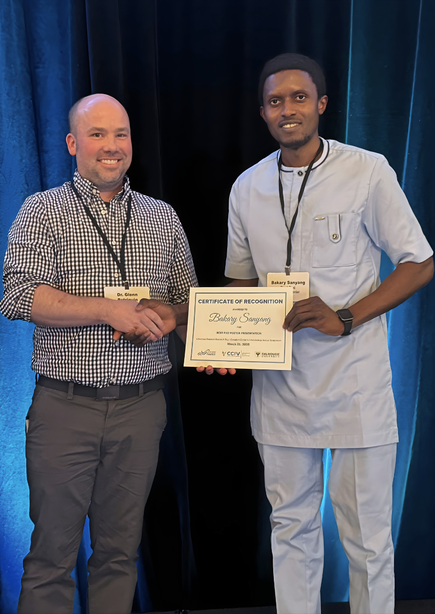 Dr. Glenn Patriquin, wearing a checkered shirt and dark pants, smiles and shakes hands with Bakary Sanyang, wearing light blue. Bakary holds the certificate for Best Poster Presenation - PhD category.