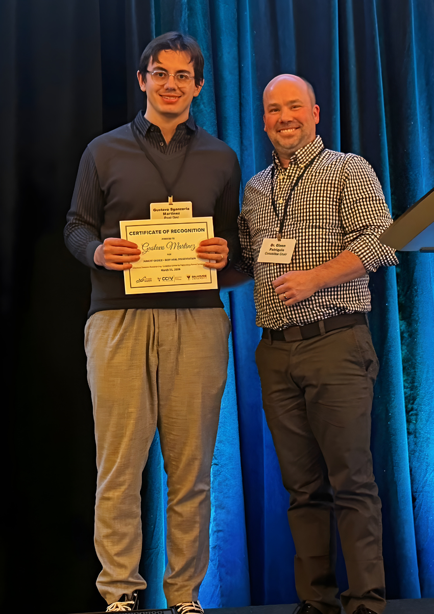 Gustavo Martinez, in a dark sweater and grey pants, holds a certificate for Judges' Choice - Best Oral Presentation. He stands next to Planning Committee Chair Glenn Patriquin who is in a checkered shirt and dark pants. Both are smiling. 