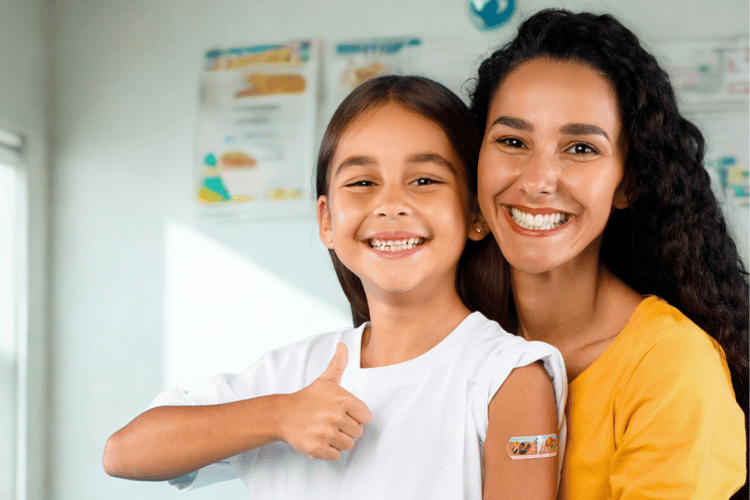 A woman smiles with her young daughter, who gives a thumbs up. They appear to be in a doctor's office. The girl has a colourful bandaid on her shoulder - she has just received a vaccine.