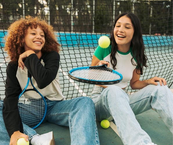 Two teen girls sit on a tennis court, smiling. The first has light brown skin and brown tightly curled hair worn loose. The other has lighter skin with straight long dark hair.