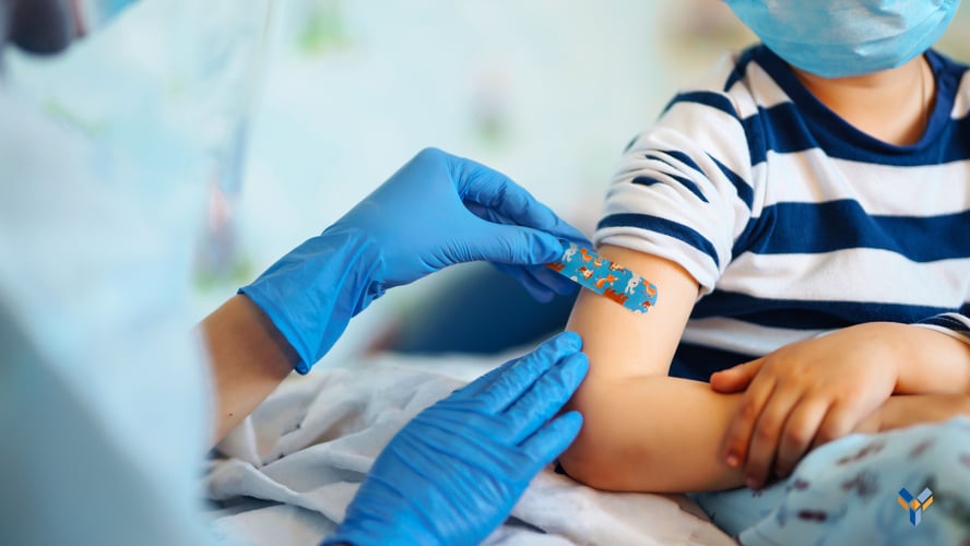 A child's arm is shown as someone wearing blue medical gloves places a blue adhesive bandage with colorful cartoon characters on it on their arm after a vaccine.