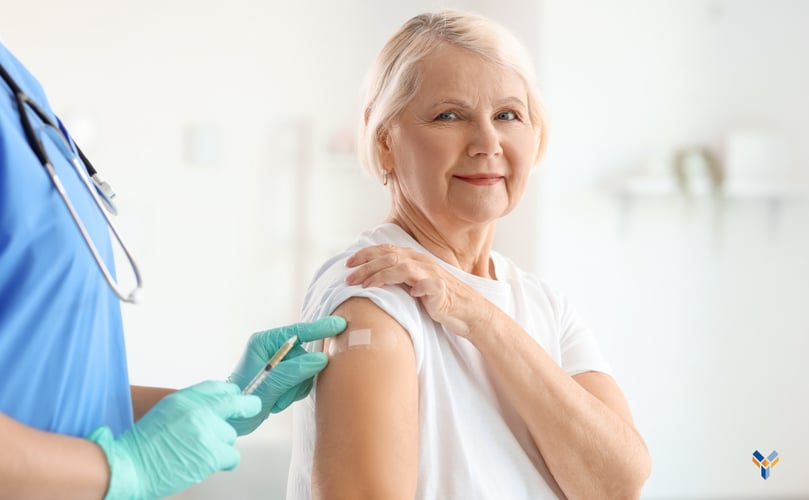 An elderly white woman with white hair smiles towards the camera, holding up a sleeve to show an adhesive bandage on her shoulder. A nurse, whose face is unseen off camera, holds a syringe in one hand as she applies the bandage with the other.